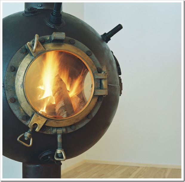 A spherical, bronze-colored metal fireplace with a circular glass window shows logs burning brightly inside. The fireplace has a rustic, industrial design with latches and bolts. It stands on a wooden floor against a plain white wall.