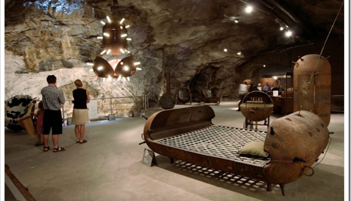 People explore a museum exhibit in a cave-like setting, featuring large, rusted, industrial sculptures and furnishings. The space is dimly lit, highlighting the rugged walls and metal artworks, including a bed frame-like structure in the foreground and various hanging pieces.