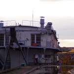 A docked, two-story houseboat with a weathered exterior, featuring metal railings and stairs leading to the upper deck. Graffiti covers part of the lower wall. A puddle reflects the boat in the foreground, and nearby are a small tree and a yellow structure. The sky is overcast.