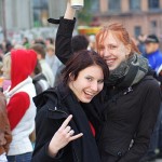 Two women smile at the camera in an outdoor crowd. One holds up a can of Luks beer and the other makes a "rock on" hand gesture. Both wear dark jackets, with one also wearing a scarf. The background includes blurred people and buildings, suggesting an event or gathering.