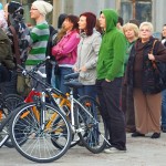 A diverse group of people stand outdoors, with some wearing casual jackets and hoodies. Bicycles are parked in the foreground. The scene appears to be a gathering or event, with individuals looking in various directions. Background shows a building facade.