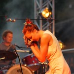 A performer in a sleeveless white outfit leans forward with both hands clasped near their face, eyes closed. In the background, a drummer plays a red drum set. Stage lights and fog create a moody atmosphere.