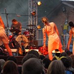 Four performers on stage: three dancers in white dresses, facing away with tilted heads, and a drummer playing intensely in the background. The stage is outdoors, surrounded by an audience. The setting has dramatic lighting with smoke effects and a brick building in the background.