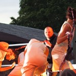 Four dancers in flowing outfits perform on an outdoor stage at dusk. They are in dynamic poses with braided hair and the soft lighting casts a warm glow. A standing man in casual attire observes the scene, and trees and a building are visible in the background.