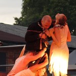 A man in a dark outfit stands entwined with a long, colorful scarf, while three women in light dresses surround him, gripping the scarf. They appear to be engaged in a dramatic performance on an outdoor stage, with trees and a building visible in the background.