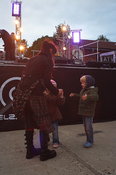 A woman in a plaid skirt and boots interacts with two children in jackets and hats in front of a stage with bright lights. The scene suggests a lively outdoor event, with the sky overcast and some people visible on stage, preparing equipment.