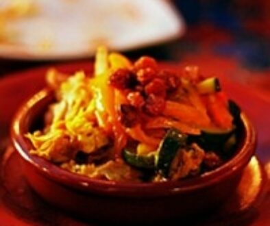 A bowl of vibrant mixed vegetables sits on a red plate. The dish includes sliced carrots, zucchini, and small pieces of roasted meat, all in a richly colored brown bowl. The setting has a warm, cozy ambiance with hints of other dishes in the blurred background.