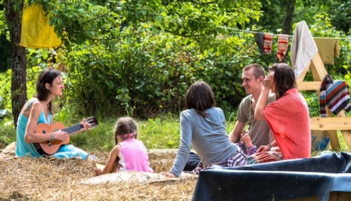 A group of five people, including a child, sit on hay in a sunny outdoor setting surrounded by trees. One person plays a ukulele. Colorful blankets hang on a line nearby. They appear relaxed and engaged in conversation, enjoying the natural environment.