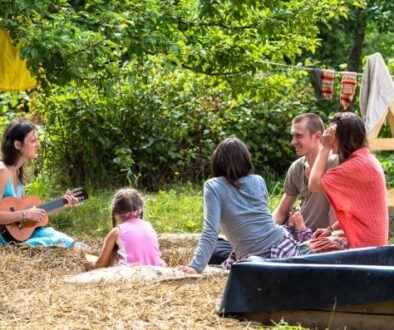 A group of five people, including a child, sit on hay in a sunny outdoor setting surrounded by trees. One person plays a ukulele. Colorful blankets hang on a line nearby. They appear relaxed and engaged in conversation, enjoying the natural environment.