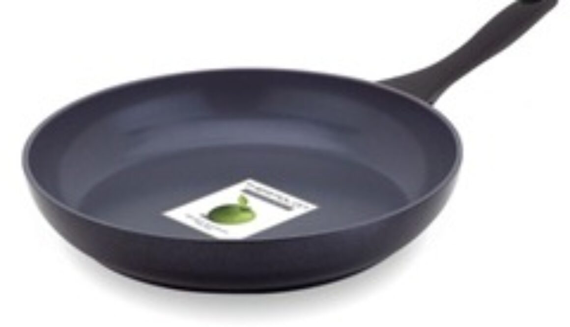 A black non-stick frying pan with a smooth matte finish is shown. It has a long, curved handle. Inside the pan, there's a partially visible label with a green apple illustration. The pan rests on a white surface.