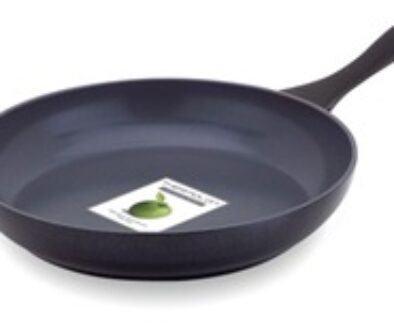 A black non-stick frying pan with a smooth matte finish is shown. It has a long, curved handle. Inside the pan, there's a partially visible label with a green apple illustration. The pan rests on a white surface.