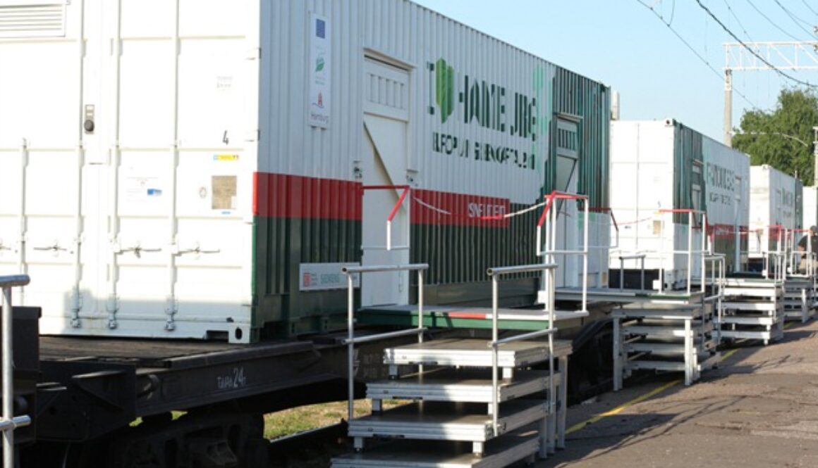 Shipping containers converted into mobile healthcare units are placed on a train flatbed. The containers are white and green with red and green stripes. Metal steps lead to the entrances, and overhead rail lines are visible above. The background includes some greenery.