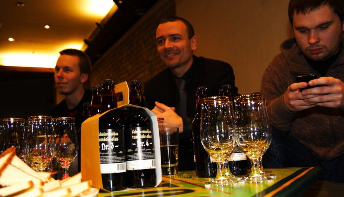 Three men sit at a table. Two are smiling and engaged, one checks his phone. On the table are bottles of dark beer in a cardboard carrier, surrounded by empty glasses and slices of bread. The setting appears warm and casual, with dim lighting and a cozy atmosphere.