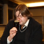 A person with shoulder-length brown hair is sitting indoors, holding a glass of dark beer close to their nose, seemingly smelling it. They are wearing a black jacket over a white shirt and black sweater. The setting appears to be a warmly lit room with large windows in the background.