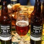 Two Valmiermuiža beer bottles flank a glass filled with dark beer on a table. The glass bears the same branding. In the background, blurred food items are visible, along with a partially filled smaller glass. The table has a yellow and green patterned surface.