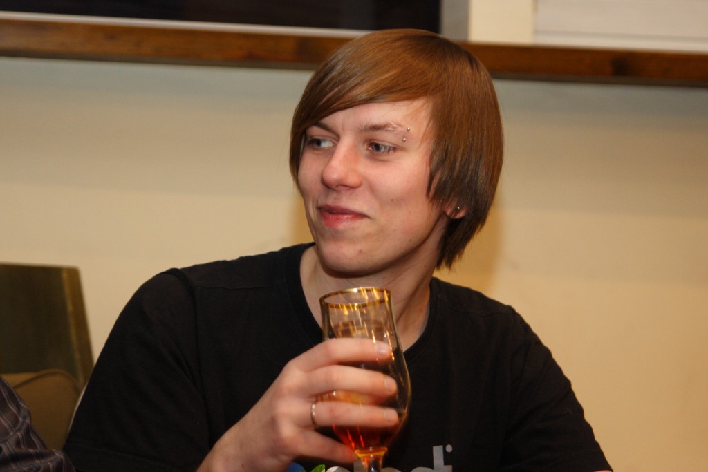 A person with straight, medium-length hair and an eyebrow piercing is sitting and holding a half-full glass of amber liquid. They are wearing a black shirt and are looking off to the side with a slight smile. The setting appears to be indoors with a neutral-colored wall and wooden trim in the background.