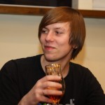 A person with straight, medium-length hair and an eyebrow piercing is sitting and holding a half-full glass of amber liquid. They are wearing a black shirt and are looking off to the side with a slight smile. The setting appears to be indoors with a neutral-colored wall and wooden trim in the background.