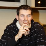 A person wearing a dark-striped hoodie sits indoors, holding a stemmed glass and taking a sip. There is a bright indoor light and a window in the background. The person has short dark hair and a light-colored scarf.