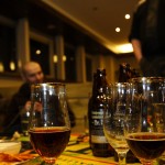 Close-up of glasses with dark beer on a table, with partially filled bottles in the background. Two people are blurred in the background, seated in a dimly lit room with warm lighting and a casual atmosphere. The table has a colorful green and yellow cloth, and some plates with food.