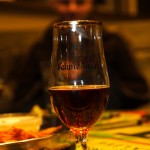 A tall glass of dark beer sits on a table adorned with colorful decorations. The glass has "Valmiermuiža" etched on it. In the blurred background, a person is seated indoors. A metal tray with food is partially visible next to the glass.