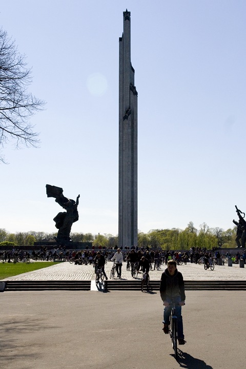 Bicycle riders leisurely pedal near a tall monument surrounded by park greenery. The monument features vertical structures with a large statue to the left. People gather at the base of the monument, enjoying the sunny day. Tree branches frame the left side under a clear blue sky.