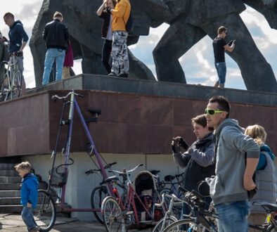 A group of people and children, some with bicycles, gather near a large, abstract sculpture. A man stands on the ledge taking a photo with a camera. Another person wears a yellow jacket, while others interact nearby. Bright, partly cloudy sky in the background.
