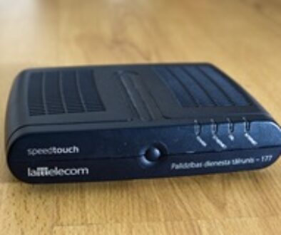 A small, rectangular black modem labeled "speedtouch" with four indicator lights on the front. It features the logo "Lattelecom" and the text "Pakalpojuma demošs iekārts – 177." The device rests on a wooden surface.