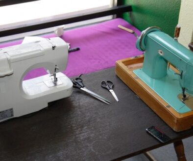 Two sewing machines on a dark table: a modern white machine on the left and a vintage turquoise machine on the right. Scissors and a mobile phone are placed between them. The background shows a window with purple fabric draped over the frame.