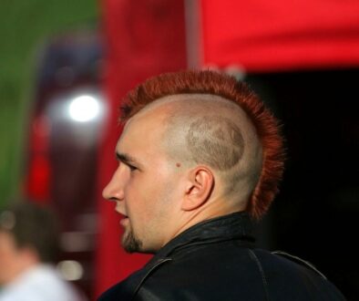 A person with a red mohawk hairstyle and a shaved pattern on the side of their head is standing outdoors. They are wearing a black leather jacket. A red structure and a blurred person in white can be seen in the background.