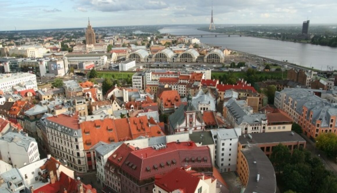 Aerial view of Riga, Latvia, showcasing a blend of colorful old town buildings with red and gray roofs. The Daugava River runs in the background, alongside modern structures, including the TV tower. The skyline features a mix of historic and contemporary architecture under a partly cloudy sky.