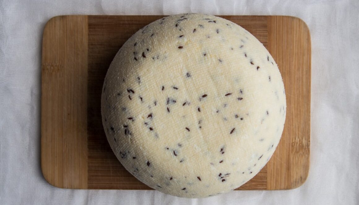 A round cheese wheel with specks of herbs or spices is placed on a rectangular wooden cutting board. The surface of the cheese has a textured, slightly rough appearance. The cutting board rests on a white cloth, creating a simple and rustic presentation.