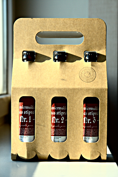 A cardboard carrier holds three glass bottles with dark caps. The bottles display labels with red and white text that include "Nr 1," "Nr 2," and "Nr 3." Sunlight casts shadows on the bottles and carrier, highlighting their shapes. The carrier has a circular stamp-like design on it.