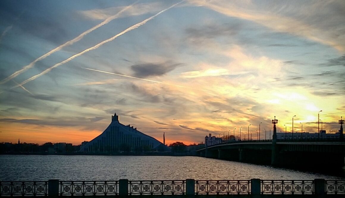 Sunset over a river with a distinctive, angular building silhouetted against the sky. A bridge spans the water to the right, and delicate clouds are streaked across the colorful sky, which transitions from orange to blue. The foreground features a railing and benches along the waterfront.
