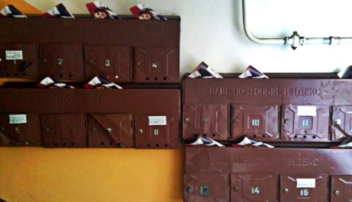 A set of brown, metal mailboxes labeled with numbers 1 to 15, mounted on a wall. Each mailbox is slightly open, overflowing with mail and flyers. The mailboxes are arranged in three rows against a background with an orange and white diagonal stripe pattern.