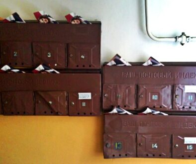 A set of brown, metal mailboxes labeled with numbers 1 to 15, mounted on a wall. Each mailbox is slightly open, overflowing with mail and flyers. The mailboxes are arranged in three rows against a background with an orange and white diagonal stripe pattern.